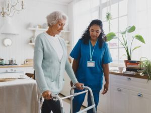 Side view of black african american nurse volunteer teaching elderly woman with gray hair in cardigan to use walker, supporting her and giving advice and instructions, standing in her kitchen